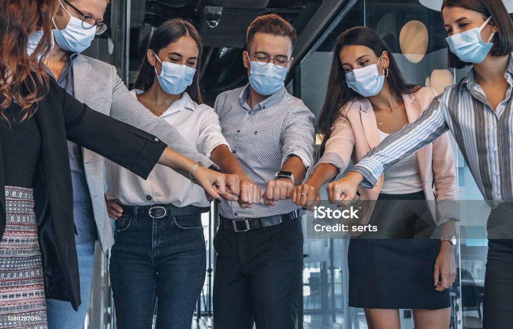 Team of colleagues with face mask joining their hands together in unity during pandemic Group of businesspeople with protective face mask joining their hands together in unity without touching  by new normal punch hand style with keeping social distance and protection for against coughs and spitting with alternative handshakes in new normal adaptation to preventing Coronavirus or Covid-19 virus spreading in a creative luxury office during New normal  viruses symptom outbreak at reopen entrance after quarantine and lockdown. Office Stock Photo