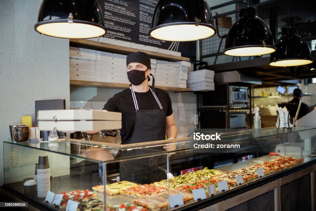 Man in protective mask and gloves passing ordered pizza in boxes over the glass counter Man working in pizza place, passing order in pizza boxes over the glass showcase counter, he is wearing protective face mask and gloves due to COVID-19 pandemic Fast Food Restaurant Stock Photo
