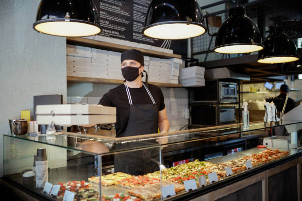 Man in protective mask and gloves passing ordered pizza in boxes over the glass counter Man working in pizza place, passing order in pizza boxes over the glass showcase counter, he is wearing protective face mask and gloves due to COVID-19 pandemic pizzeria stock pictures, royalty-free photos & images