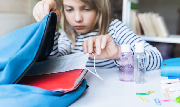 Little girl packing blue backpack in kids's room. Face mask, bottle of sanitizer, stationery, pens, multicolored markers, notebooks. Back to school. Mom's hygiene,safety precautions after coronavirus. niña empacando mochila azul en la habitación de los niños. máscara facial, botella de desinfectante, papelería, bolígrafos, marcadores multicolores, cuadernos. regreso a la escuela. higiene de la madre, precauciones de seguridad después del coronavi