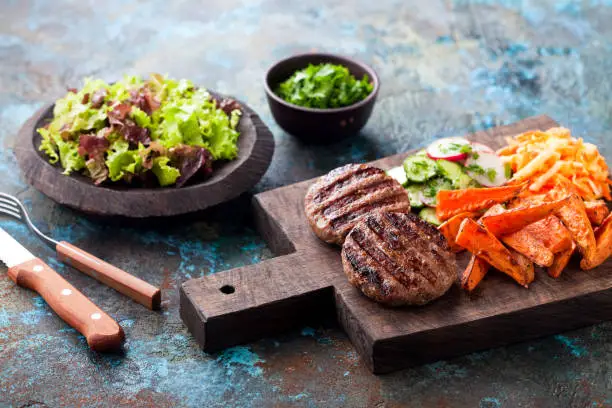 Grilled burger cutlets with baked sweet potato, vegetables and salad served on wood chopping board, selective focus