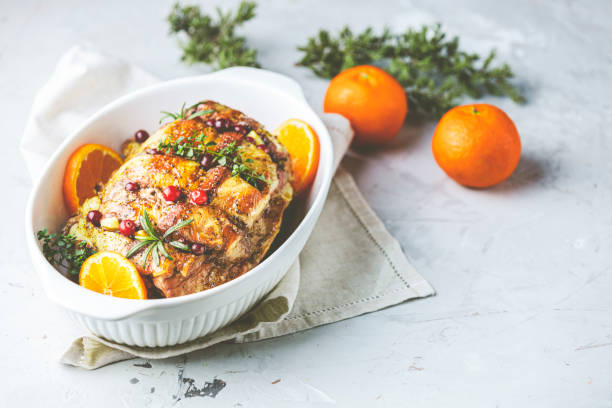Roasted pork in white dish, christmas baked ham with cranberries, tangerines, thyme, rosemary, garlic on light table surface. stock photo