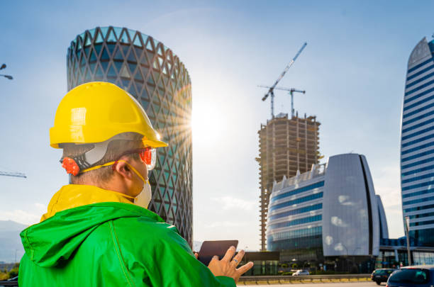 istock Young engineer with yellow hardhat,  N95 protective mask  and green protective workwear using digital tablet in front of modern business buildings and construction site, lens flare 1280300367