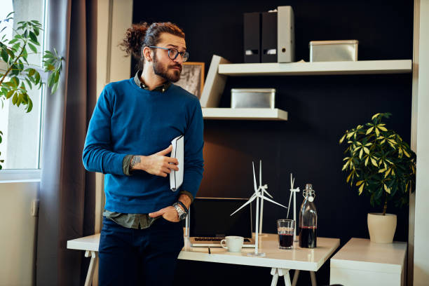 istock Young serious handsome hipster standing in his office, holding documents under armpit and preparing for important meeting with his boss. Sustainable development concept. 1280307382