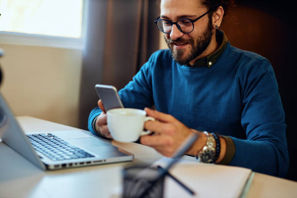 istock Smiling handsome bearded hipster sitting in his office, drinking coffee and typing a message on the phone on a break. He is hanging on social media sites. 1280307422