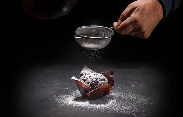 View of the hand of the pastry chef putting powdered sugar on delicious chocolate muffin on dark background