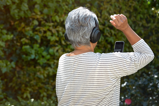 Rear view of happy senior woman dancing and listening to music with headphones on a sunny day in the garden