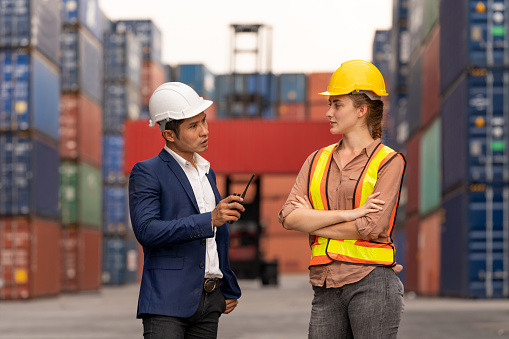 Technician consult with worker about transport cargo container in shipping yard. Businessmen and engineer wears hard hat standing in front of Reach stacker vehicle. Logistic and transportation concept