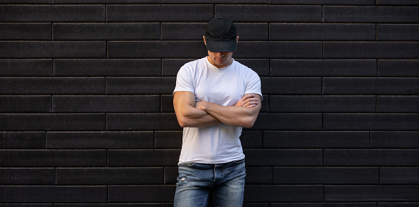 Man in white t-shirt standing against black brick wall with arms crossed