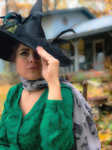 Beautiful woman dressed as a wait here for Halloween with her face peeking out from a large black witch hat. She is sitting in an old wooden chair outside of her home.