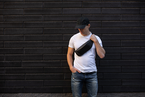 Mockup of man in white t-shirt standing against brick wall holding black waist bag