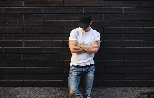 Man in white t-shirt standing against black brick wall with arms crossed