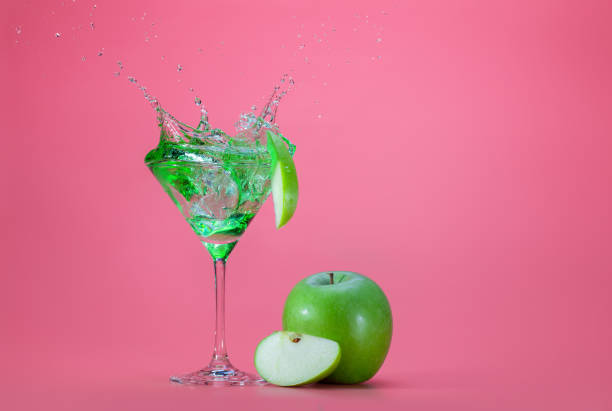 パステルピンクの背景にマティーニグラスから飛び出す緑のカクテル - apple martini ストックフォトと画像