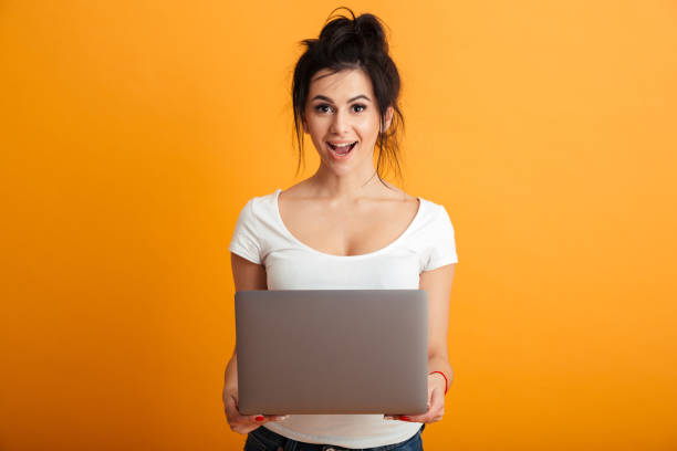 istock Gorgeous woman 20s with brown hair in bun looking on camera while working or communicating on silver laptop, over yellow background 1280348618