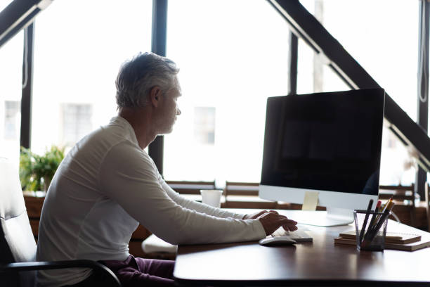 istock Casual Grey-haired Mature Man Working At Desk In Busy Creative Office. 1280348787