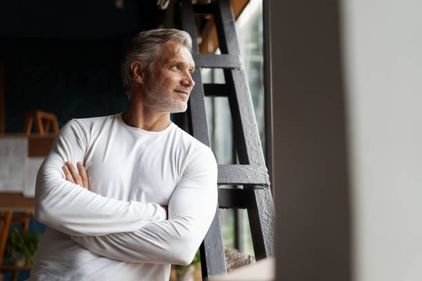 Business lifestyle. Casual Grey-haired Mature professional handsome businessman standing near window in his office Business lifestyle. Casual Grey-haired Mature professional handsome businessman standing near window in his office while thinking about his goals handsome people stock pictures, royalty-free photos & images