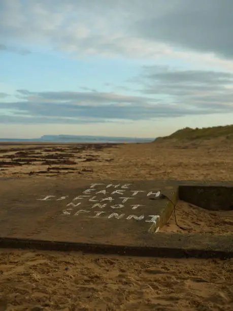 Bleak and desolate image of Redcar Beach, showing a derelict World War Two defensive structure bearing an anti-science slogan in the foreground. Mid-ground and background are partly defocused beach and sand-dunes, with a partly clouded blue winter sky.