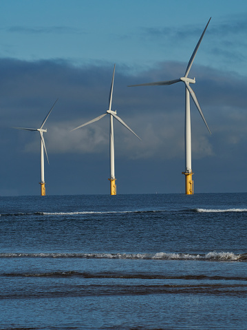 Image of a trio of near-shore wind turbines, lit by sharp winter sun, with a background of looming, threatening clouds and a foreground of sea and breaking waves.