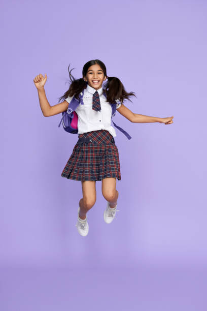 entusiasta divertente ragazza della scuola elementare indiana con borsa a zaino con l'uniforme che salta volando isolata su sfondo viola. felice pazzo latino bambino studentessa studentessa sensazione di libertà divertendosi. - schoolgirl school children isolated child foto e immagini stock