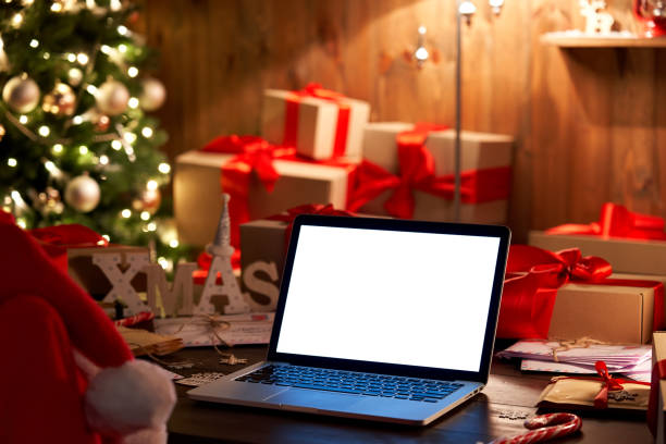 Laptop computer with white blank empty mock up screen on Merry Christmas table with presents gifts boxes, decorated Xmas tree in cozy Santa house background. Ecommerce website online shopping delivery