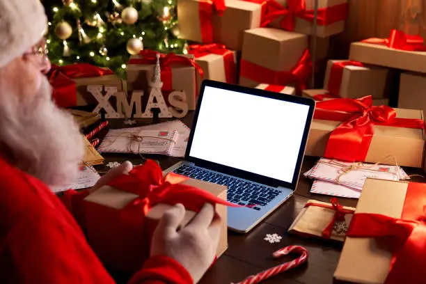 Photo of Over shoulder view of Santa Claus holding xmas gift using laptop computer with white blank empty mock up screen monitor for e commerce website ad sitting at decorated table on Merry Christmas eve.