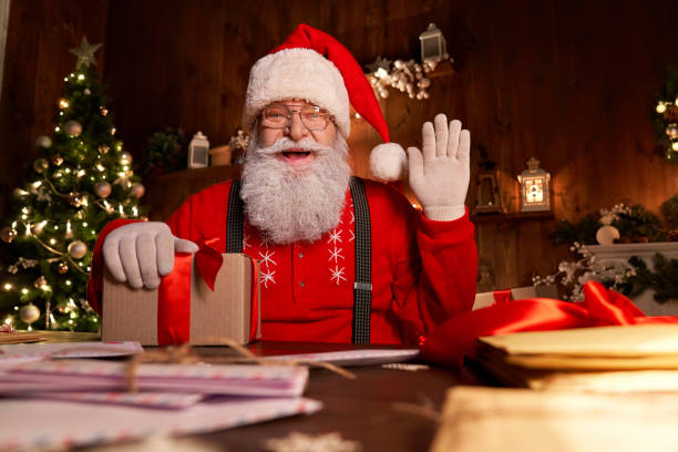 glücklicher weihnachtsmann, heiliger nikolaus hält brief mit blick auf die kamera, sprechen mit webcam-videoanruf oder aufnahme frohes neues jahr, frohe weihnachtsgrüße am weihnachtsabend sitzen am tisch spät zu hause. - grüßen fotos stock-fotos und bilder