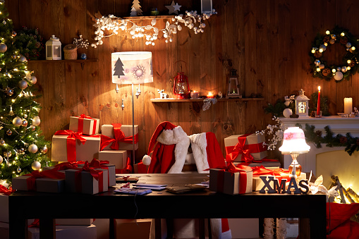 Traje de Papá Noel y sombrero colgado en la silla en la mesa con regalos de decoración Feliz Navidad regalos regalos en la víspera de vacaciones en el acogedor interior del taller de Santa Casa tarde en la noche con luz en el árbol de Navidad y chimene photo