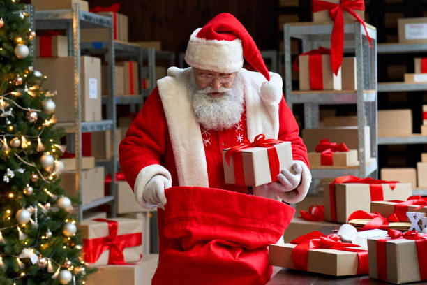 istock Old funny Santa Claus, Saint Nicholas packing presents gift boxes in sack bag preparing post shipping fast xmas delivery parcels standing in workshop. Merry Christmas shipping delivery concept. 1280356918