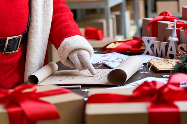 Santa Claus wearing costume reading parchment roll letter wish list standing at table with presents in workshop preparing for holiday on xmas eve. Merry Christmas wishlist background concept, closeup. papá noel usando una lista de deseos de letra de pergamino de lectura de disfraces de pie en la mesa con regalos en el taller preparándose para las vacaciones en la víspera de navidad. feliz navidad concepto de fondo de la lista de deseos, primer plano.