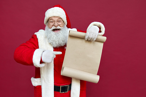 Feliz excitado viejo Bar Claus barbudo que lleva traje sosteniendo Rollo de papel de la lista de deseos de Feliz Navidad apuntando con el dedo a la letra vacía en blanco lista de deseos de Navidad de pie aislado en el fondo rojo, copiar el espacio. photo
