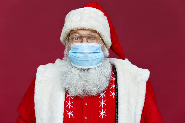 retrato de cerca de la vieja barba divertida sorprendió a santa claus usando traje, gafas, máscara facial mirando a la cámara de pie en el fondo rojo de navidad. concepto de protección de seguridad de coronavirus covid 19. - santa hat christmas hat headwear fotografías e imágenes de stock