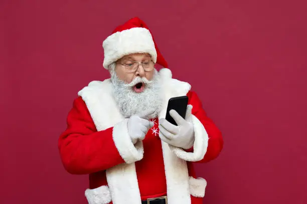 Photo of Surprised funny amazed old bearded Santa Claus wearing costume holding cell phone using mobile app on smartphone shocked by Christmas promotion, xmas applications ads isolated on red background.