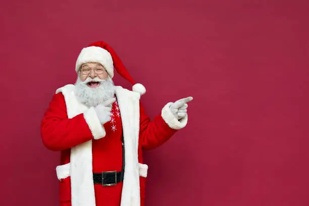 Photo of Funny happy excited old bearded Santa Claus face wearing costume looking at camera showing pointing fingers aside advertising Christmas promotion, New Year xmas discount ad isolated on red background.