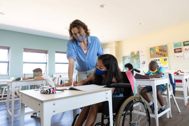 istock Disable girl wearing face mask writing while sitting on wheelchair in class 1280360731