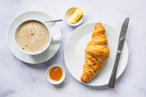 A freshly baked French croissant is sitting on a white side plate. There is a pot of butter and jam beside the croissant and a cup and saucer of freshly brewed coffee. The background is white marble.