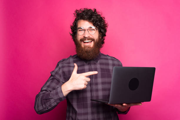 istock Young bearded man with glasses is smiling at the camera holding a computer and pointing at it near a pink wall 1280367654