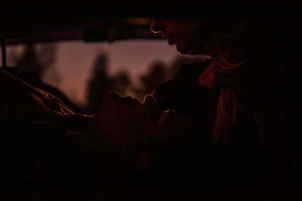 silhouette der liebhaber im auto auf dem rücksitz. liebespaar zog sich in der dämmerung zurück, um den abend zusammen zu verbringen. das mädchen beugte sich, um den kerl zu küssen, der unten liegt und in ihre augen schaut. leidenschaft - passion kissing black white stock-fotos und bilder
