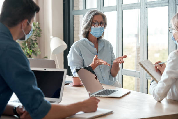 old female mentor coach wearing face mask training young workers at group meeting in reopen office. middle aged businesswoman teacher working with students at university class. social distance concept - women professor mature adult human face imagens e fotografias de stock