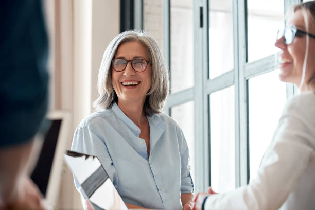 istock Happy mature old female mentor coach supervisor training young interns at group office meeting professional workshop. Smiling middle aged teacher professor laughing with students at university class 1280371018
