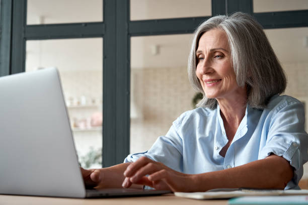 istock Smiling stylish mature middle aged business woman using laptop computer sitting at workplace desk. Happy senior older lady, 60s grey-haired businesswoman working typing on pc at home or from office. 1280371026