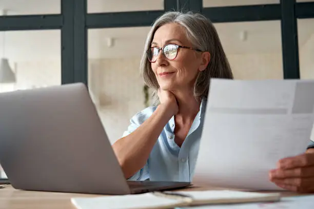 Photo of Smiling mature middle aged business woman using laptop working on computer sitting at desk. Happy old businesswoman hr holding cv interviewing distance applicant, senior seeker searching job online.