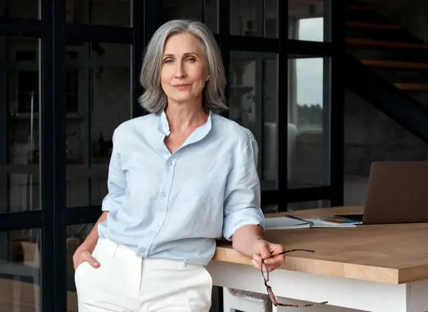 Photo of Confident stylish european mature middle aged woman standing at workplace. Stylish older senior businesswoman, 60s gray-haired lady executive leader manager looking at camera in office, portrait.