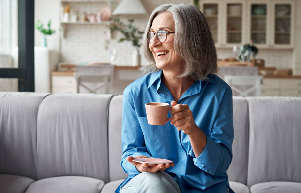 glücklich schöne entspannte ältere erwachsene grauhaarige frau trinken kaffee entspannend auf dem sofa zu hause. lächelnde stilvolle mittelalter 60er jahre dame genießen ruhe auf der couch im modernen wohnzimmer. - female healthy lifestyle wellbeing elegance stock-fotos und bilder