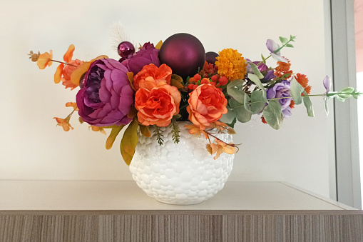 Artificial flowers in a white vase on a shelf in the room. Fabric flowers as an element of interior decor. Artificial peonies, a bouquet of flowers in a round vase.