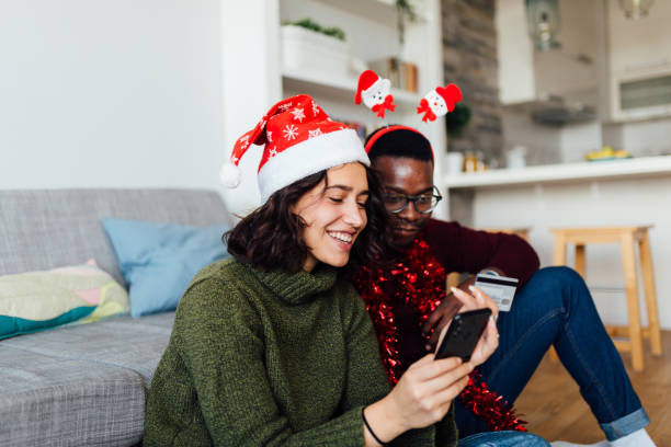Happy interracial couple shopping from home with smart phone and a credit card Joyful young interracial couple having fun at home during winter holidays, shopping for gifts using a smart phone and a credit card, smiling holiday shopping stock pictures, royalty-free photos & images