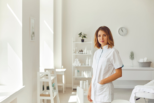 Young smiling woman cosmetologist or dermatologist standing and looking at camera in light beauty spa salon over cosmetics at background. Cosmetology and skincare concept