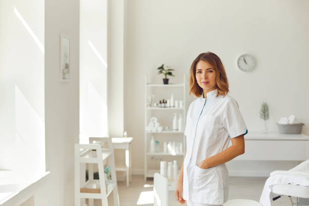 Smiling woman cosmetologist or dermatologist standing and looking at camera in beauty spa salon Young smiling woman cosmetologist or dermatologist standing and looking at camera in light beauty spa salon over cosmetics at background. Cosmetology and skincare concept beautician stock pictures, royalty-free photos & images
