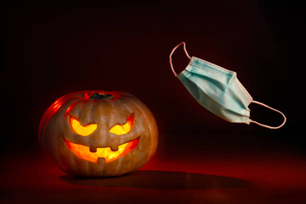 istock Illuminated halloween pumpkin with devilish face and coronavirus protective surgical mask flying on one side. New normal 1280396309