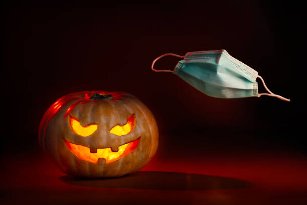 istock Illuminated halloween pumpkin with devilish face and coronavirus protective surgical mask flying on one side. New normal 1280396317