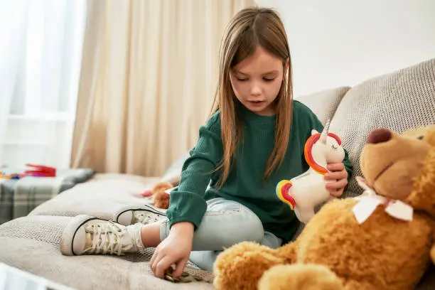 The girl is sitting alone on a sofa in a large guestroom counting coins and playing with a unicorn moneybox, having her teddybear nearby, child playing at home
